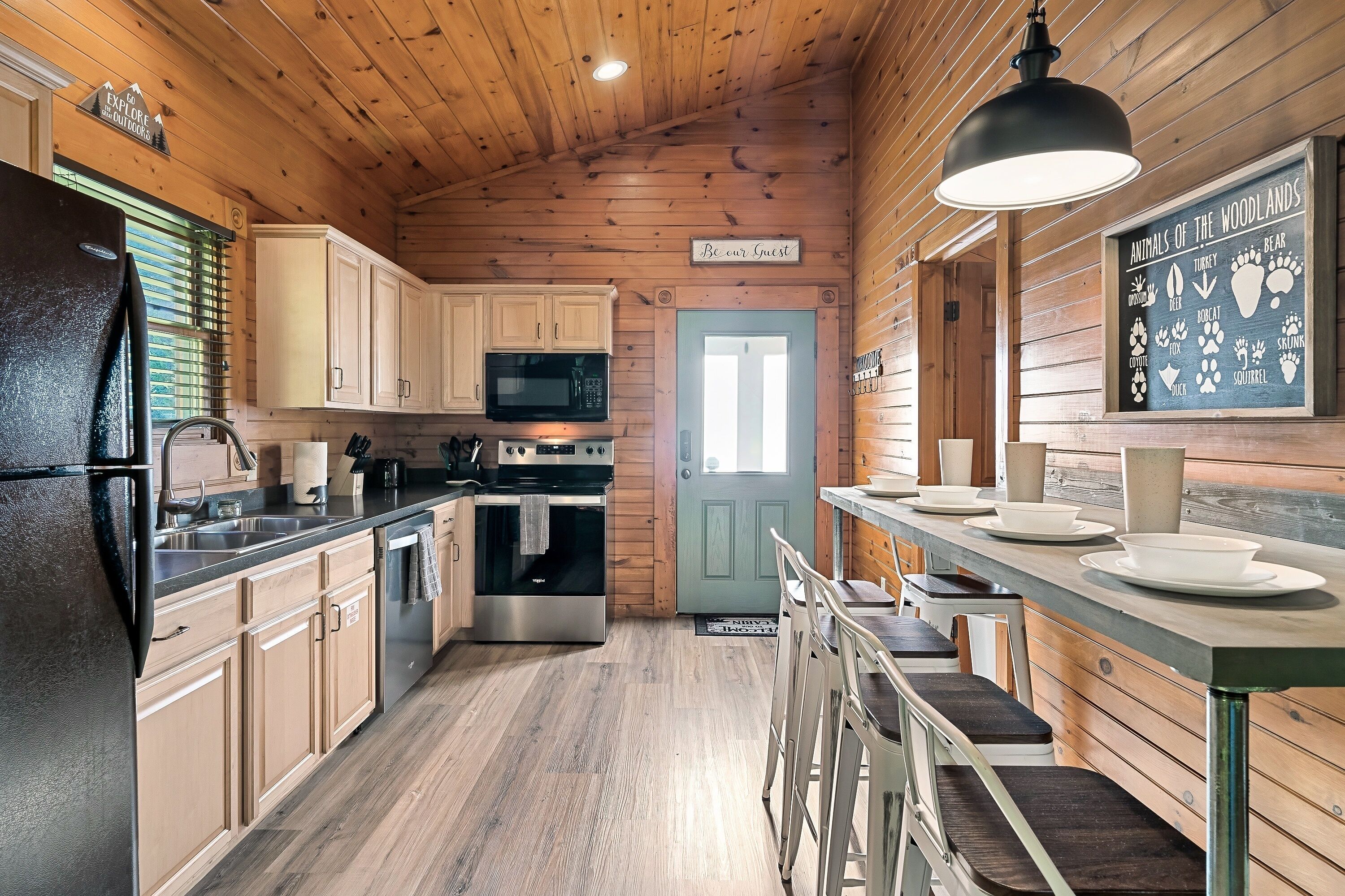 Une cuisine de style rustique, avec des éléments en bois, un réfrigérateur noir, des appareils en acier inoxydable et un tableau noir servant de menu, fixé au mur.