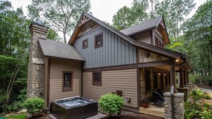 A two-story log cabin with a hot tub, stone chimney, and a covered porch.