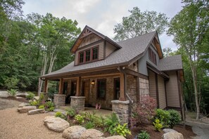 Une cabane en rondins à deux étages, avec des fondations en pierre, une véranda qui fait le tour de la maison et un paysage boisé en arrière-plan.