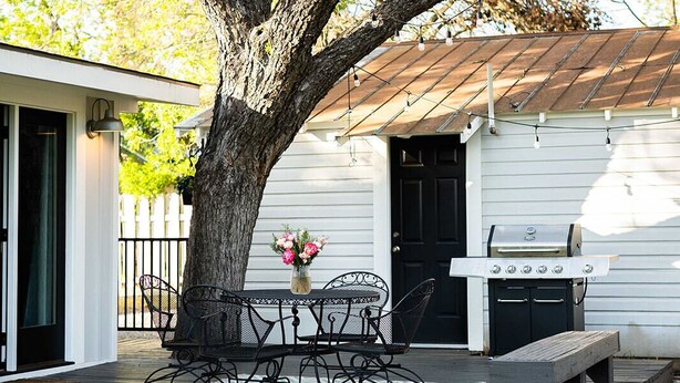 A backyard with a large tree, a grill, and a patio with metal chairs and a table.