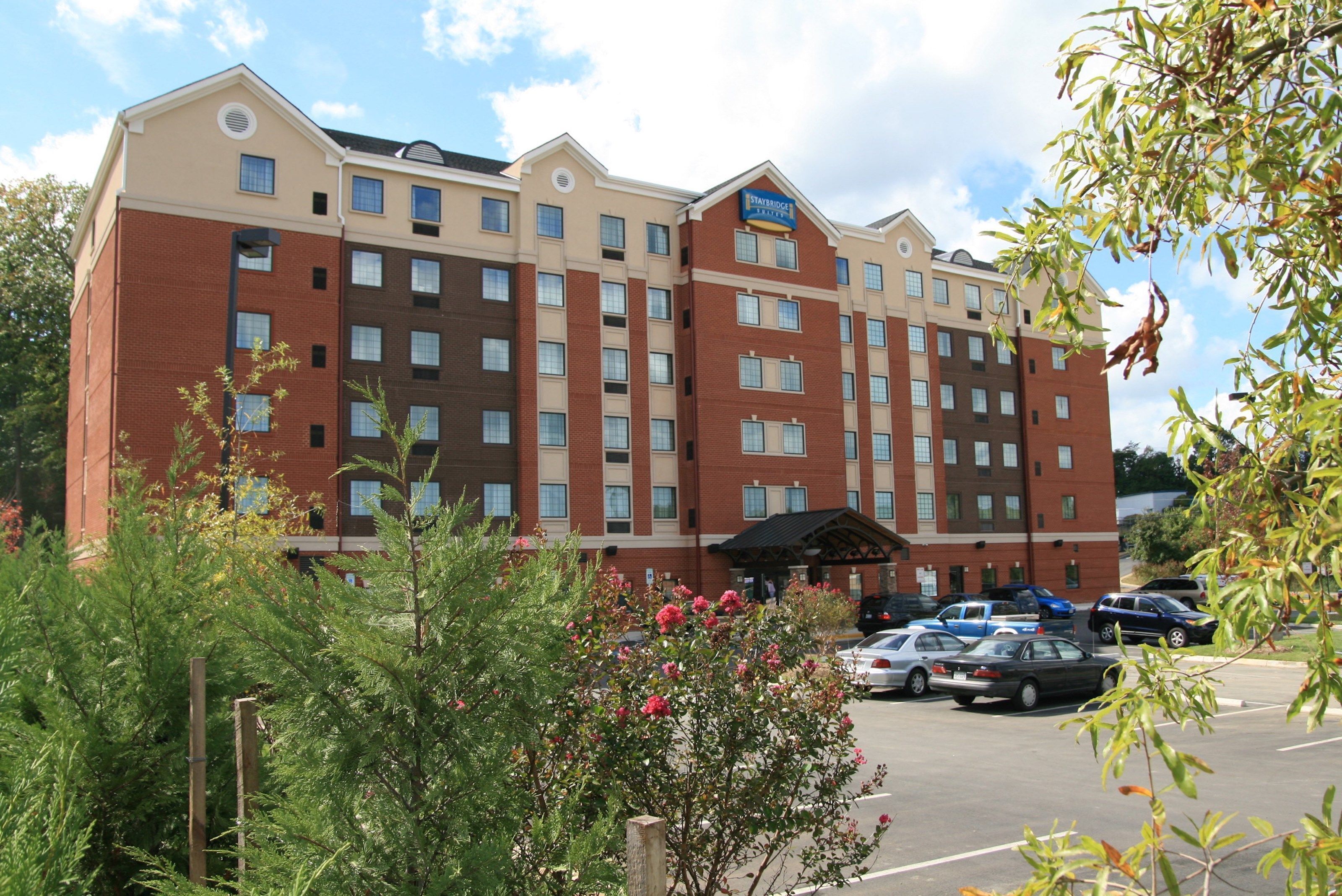 A multi-story hotel with a brick facade, a central entrance, and a parking lot with several cars.