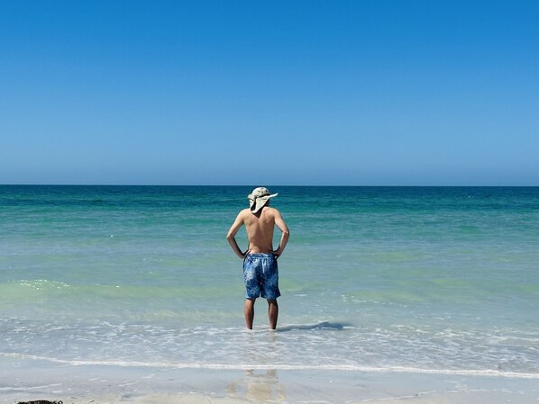 Une personne se tient sur une plage, l’eau est claire et le ciel est bleu.