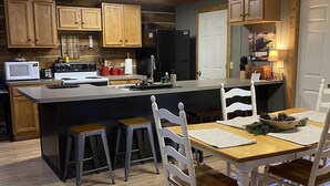 Una cocina con armarios de madera, un refrigerador negro, un microondas y un comedor con una mesa de madera y sillas blancas.