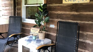 Una cabaña con interior de madera, dos sillas negras, una mesa con mantel a cuadros, un cartel de bienvenida y un letrero que dice "CABIN BY THE LAKE".