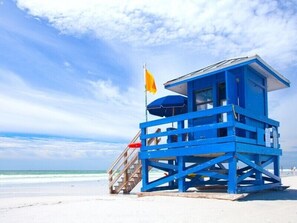 A blue lifeguard tower with a flag on top, located on a beach with clear skies and ocean in the background.
