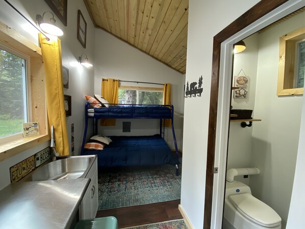 A compact room with bunk beds, a small kitchenette, a window with curtains, and a bathroom with a toilet and sink. - Cute & Cozy Tiny House in the Woods (Seward)