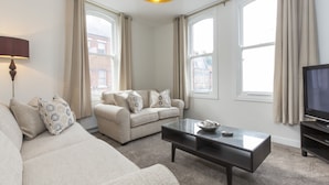 A living room with a sofa, a coffee table, a television, and large windows with curtains.