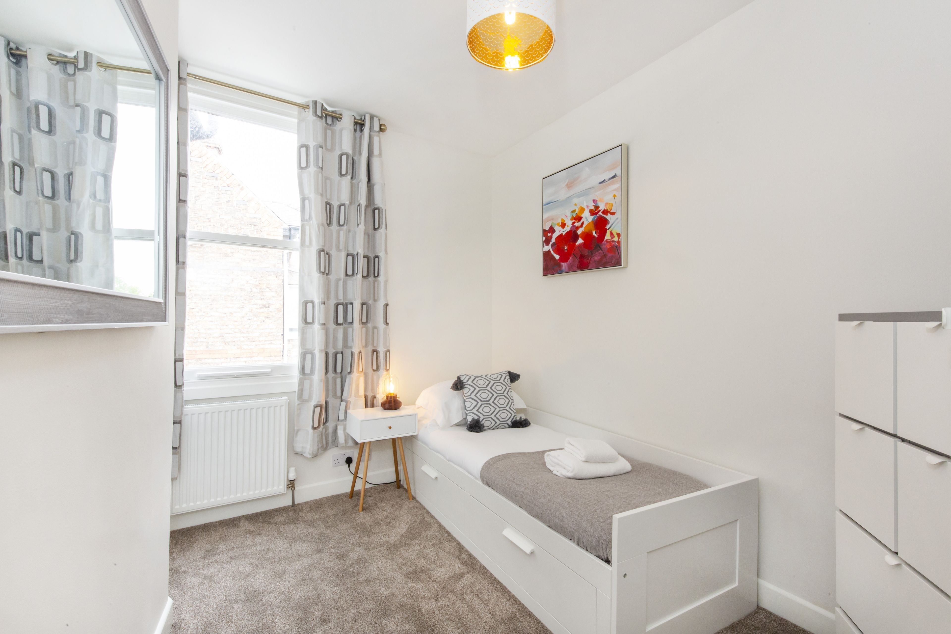 Een kleine, witte slaapkamer met een enkel bed, een nachtkastje, een radiator en een raam met gordijnen.