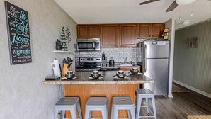 A kitchen with wooden cabinets, a stainless steel refrigerator, a microwave, and a coffee maker. There are four stools at the counter and a ceiling fan above.