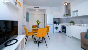 Een moderne keuken met witte kastjes, een ingebouwde oven en een magnetron. Er is een eethoek met een glazen tafel en gele stoelen.