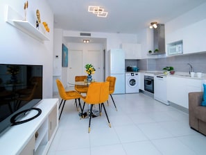 A modern kitchen with white cabinets, a built-in oven, and a microwave. There is a dining area with a glass table and yellow chairs.