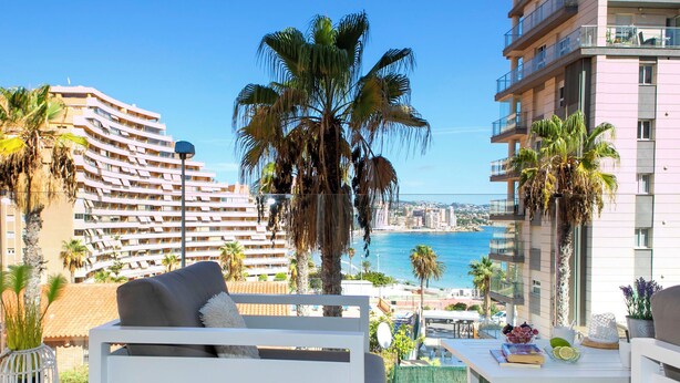 Een balkon met een bank, een tafel met fruit en boeken, en uitzicht op zee en gebouwen.