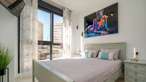 Een slaapkamer met een groot bed, een nachtkastje, een tafeltje met een lamp, een raam met dunne gordijnen en een schilderij aan de muur.