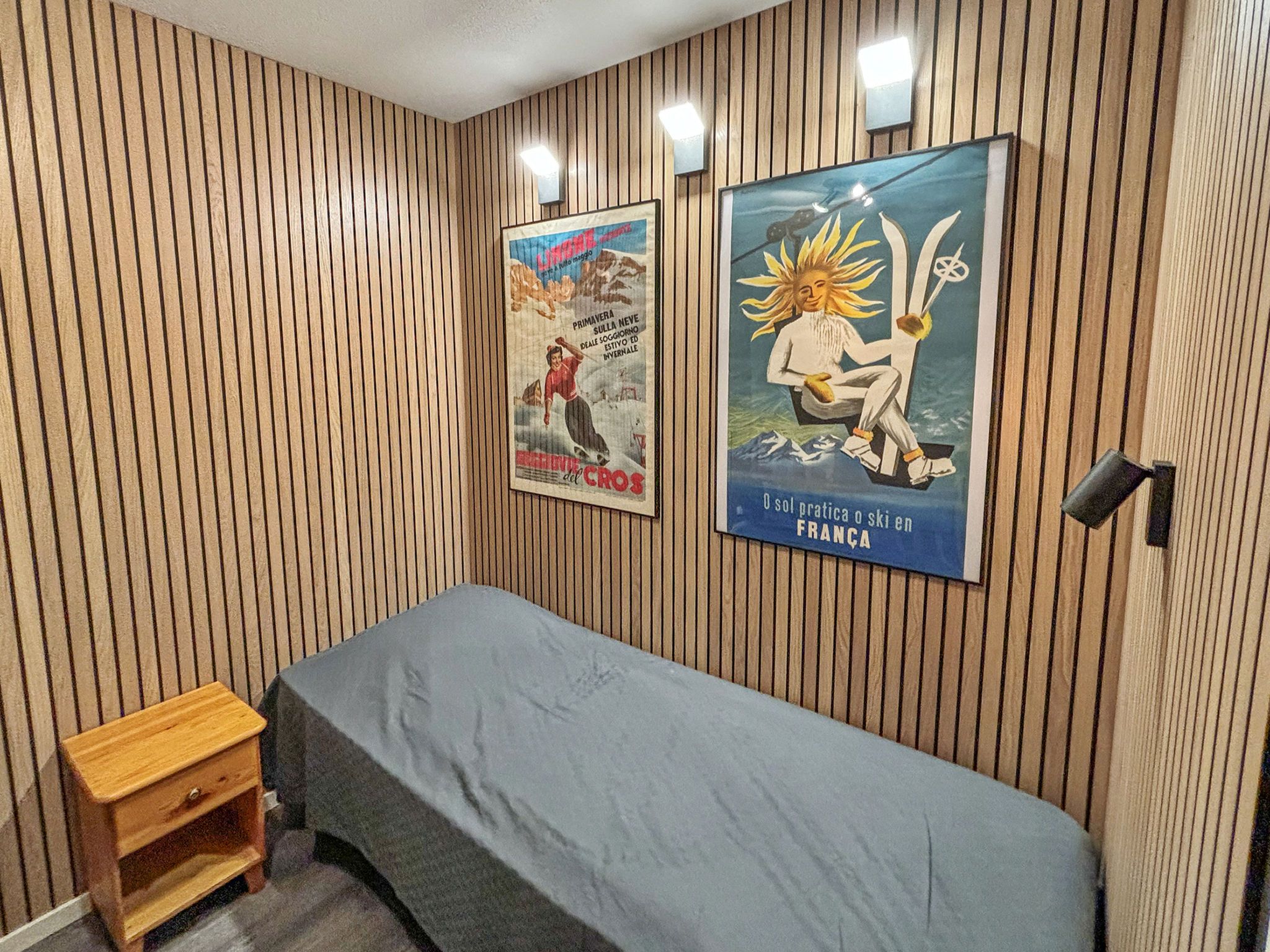 Une pièce aux murs recouverts de lambris en bois, un lit avec un matelas gris et deux affiches accrochées au mur.