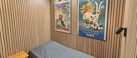 Une pièce aux murs recouverts de lambris en bois, un lit avec un matelas gris et deux affiches accrochées au mur.