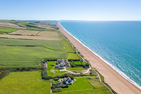 An aerial view of a coastal property with a beach, green fields, and a large house with multiple rooms.