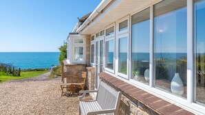 A coastal property with a patio area featuring a bench and chairs, large windows offering ocean views, and a stone wall.