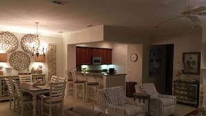Amplia sala de estar con un sofá grande, sillones estampados y una zona de comedor con una mesa y sillas de madera. Se observa una cocina con armarios rojos y un microondas al fondo.