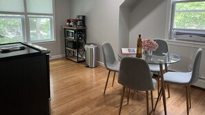 A dining area with a round glass table, grey chairs, a small trash bin, a coffee maker, and a microwave on a black metal shelf. - Great Comfortable Private Studio (Barnstable)