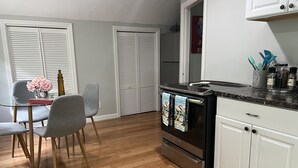 A modern kitchen with a black stove, white cabinets, and a dining area with grey chairs and a glass table. - Great Comfortable Private Studio (Barnstable)