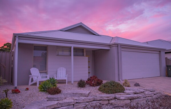A modern house with a covered porch, two white chairs, and a variety of small plants and rocks in the garden.