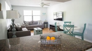 Une cuisine moderne avec un comptoir en granit, un coin repas avec des chaises de couleur turquoise, un canapé gris agrémenté de coussins à motifs et un ventilateur de plafond.