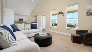 A spacious living room with a sectional sofa, ottoman, and wicker furniture. A kitchen is visible in the background with white cabinets and a microwave. - 3 BR Beach view -For the Good Times Beach House (Galveston)