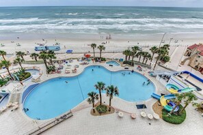A large swimming pool with a water slide, surrounded by palm trees and beach chairs. - Catch n` Relax - Ocean Walk (Daytona Beach)
