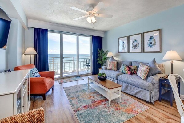 A spacious living room with a large window, a ceiling fan, a sofa, a coffee table, and a TV. - Catch n` Relax - Ocean Walk (Daytona Beach)