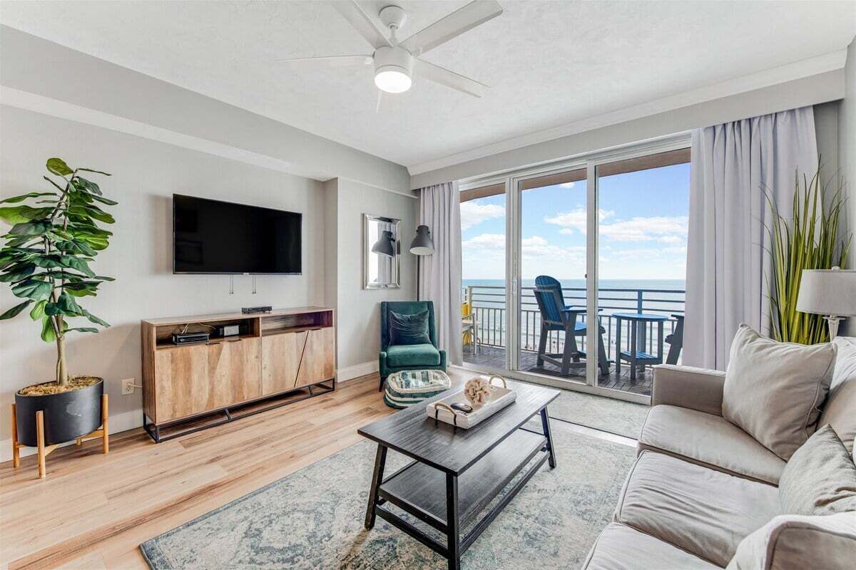 Un salon moderne doté d’un téléviseur à écran plat, d’un meuble en bois, d’un fauteuil vert, d’un canapé beige et d’un balcon offrant une vue sur l’océan.