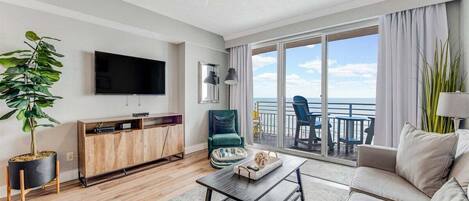 Un salon moderne doté d’un téléviseur à écran plat, d’un meuble en bois, d’un fauteuil vert, d’un canapé beige et d’un balcon offrant une vue sur l’océan.