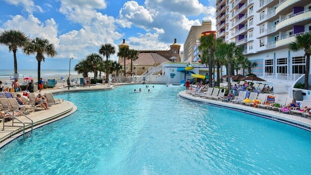 A large pool with lounge chairs and palm trees, surrounded by a hotel building with balconies. - Lattitude Adjustment - Ocean Walk (Daytona Beach)