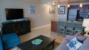 A living room with a flat-screen TV, a coffee table with a turtle sculpture, a sofa with a crab pillow, and a kitchen area with a refrigerator and a sink. - Lattitude Adjustment - Ocean Walk (Daytona Beach)