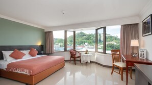 Une chambre d’hôtel avec un grand lit, un bureau, une chaise et une petite table. De grandes fenêtres offrent une vue sur l’extérieur.