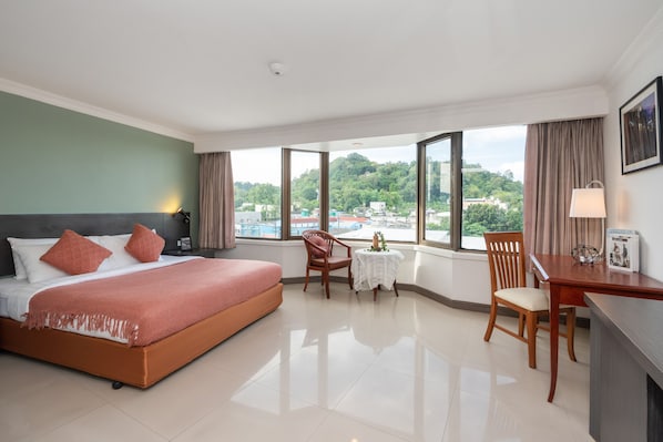 A hotel room with a large bed, a desk, a chair, and a small table. There is a view of the outside through large windows.