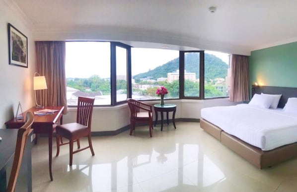 Habitación de hotel con una cama grande, un escritorio con silla, una mesita con un florero, y ventanas amplias que ofrecen vistas a la ciudad y las colinas.
