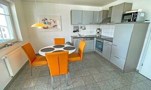 A modern kitchen with grey cabinets, a built-in oven, and a microwave. There is a dining table with orange chairs and a hanging light fixture. - 04 Ferienwohnung Greta mit Balkon - Appartementhaus Greta im Ostseebad (Sellin)