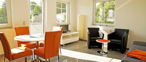 A modern living room with a dining area, featuring a round table, orange chairs, a grey sofa, and a TV on a white cabinet.