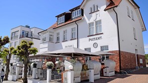 A white building with a sign reading "Postamt" and a clock. There are outdoor seating areas with umbrellas and wicker furniture. - 05 Ferienwohnung Greta mit Balkon - Appartementhaus Greta im Ostseebad (Sellin)