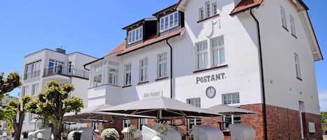 A white building with a sign reading "Postamt" and a clock. There are outdoor seating areas with umbrellas and wicker furniture.