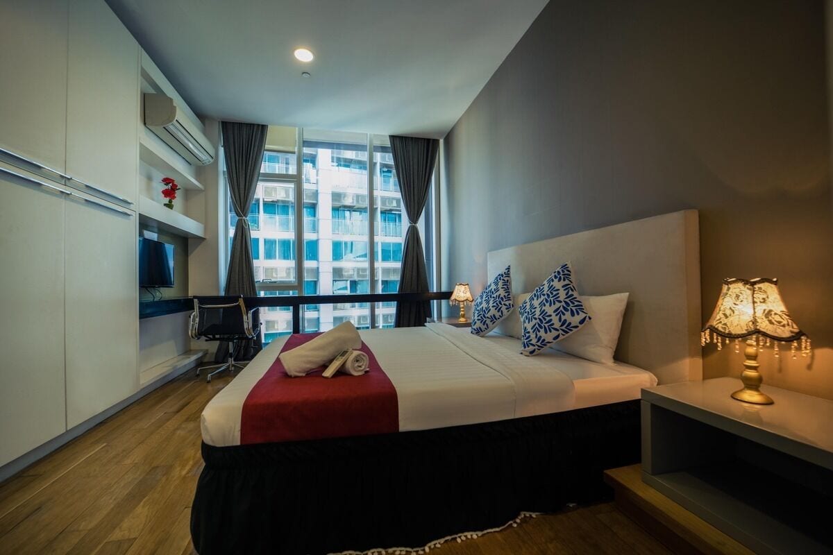 A hotel room with a bed, bedside table, lamp, and a view of buildings through large windows.