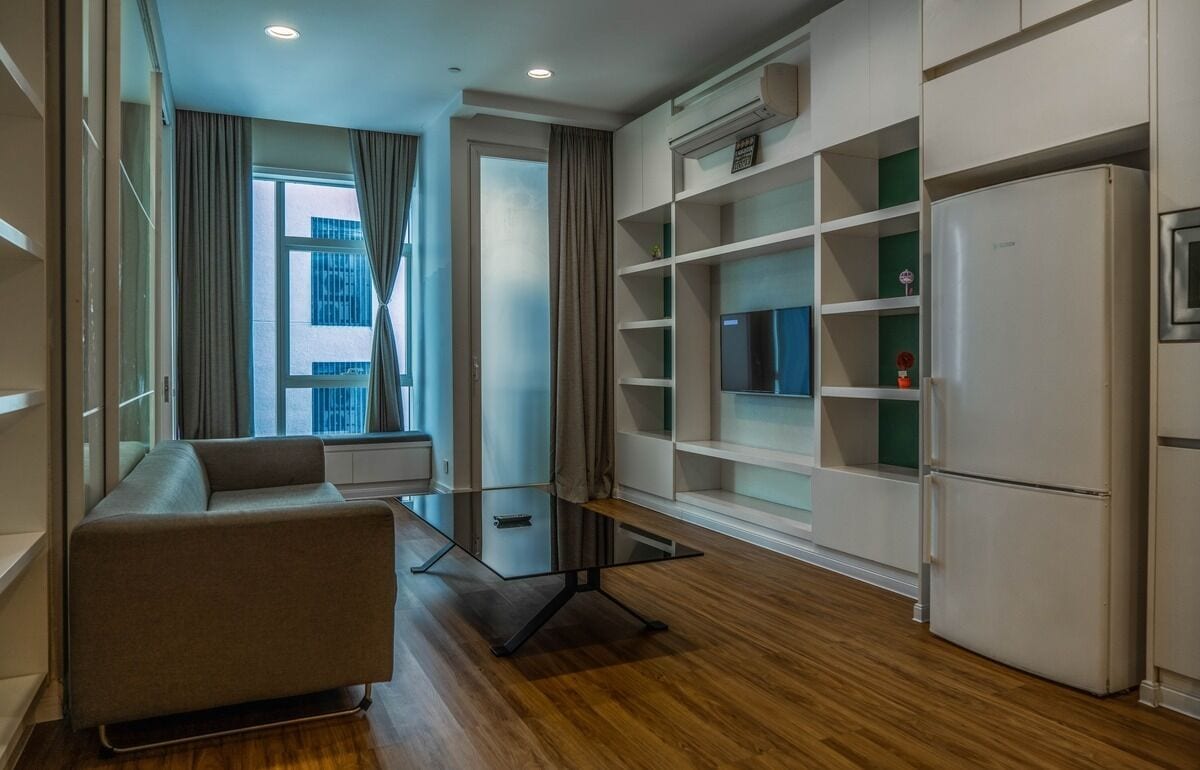 A modern apartment with a glass table, a sofa, built-in shelves, a refrigerator, and a window with curtains.