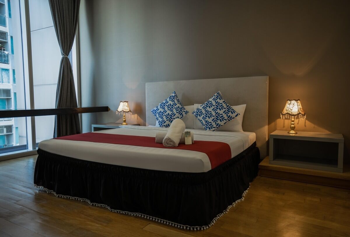 A hotel room with a large bed, two bedside lamps, a red throw blanket, and a view of a building through a window.