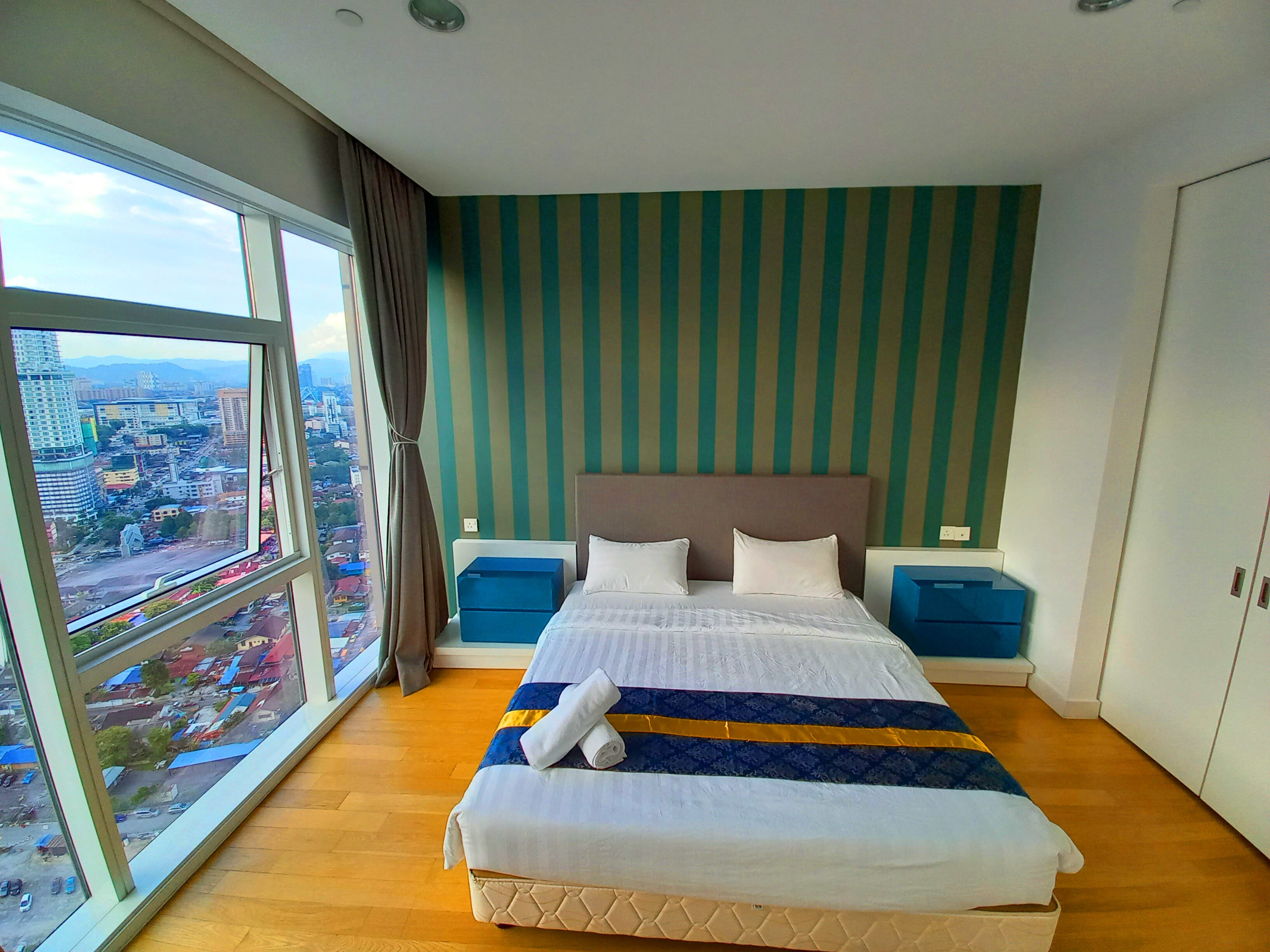 Une chambre d’hôtel avec une grande fenêtre, un lit recouvert d’une courtepointe à rayures bleues et blanches, et une vue sur le paysage urbain.