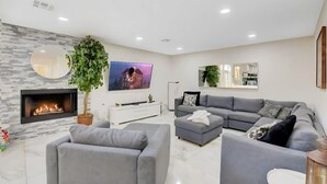 Een moderne woonkamer met een open haard, grijze banken, een flatscreen televisie en een grote spiegel.
