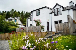 A white two-story house with a wooden fence, a front door, and a staircase leading to the entrance. - A home in the Scottish Highlands. (Dulnain Bridge)