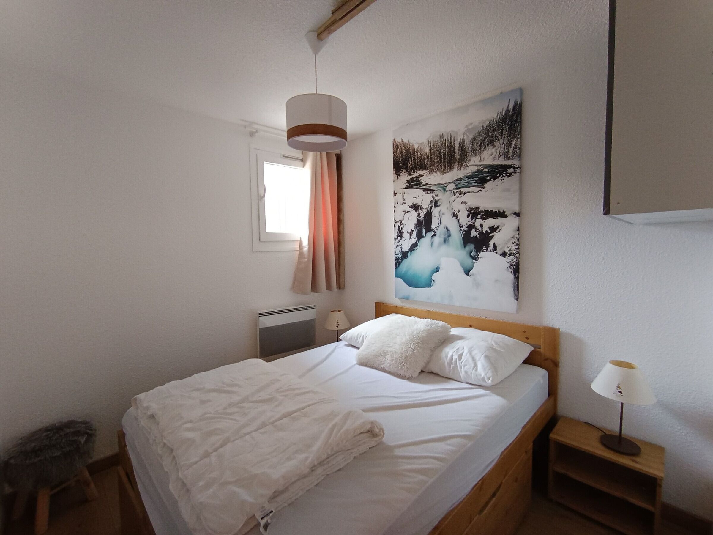 Een net opgemaakt bed met witte lakens en kussens, een bedlamp, een radiator en een schilderij van een besneeerd landschap aan de muur.