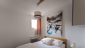 Una cama bien hecha con sábanas y almohadas blancas, una lámpara de noche, un radiador y un cuadro de un paisaje nevado en la pared.