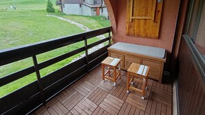 Un balcón con banco y mesa de madera, una puerta de madera y vista a un campo verde y edificios al fondo.