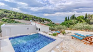Una piscina en la azotea con vistas al mar, rodeada de vegetación y un edificio blanco.
